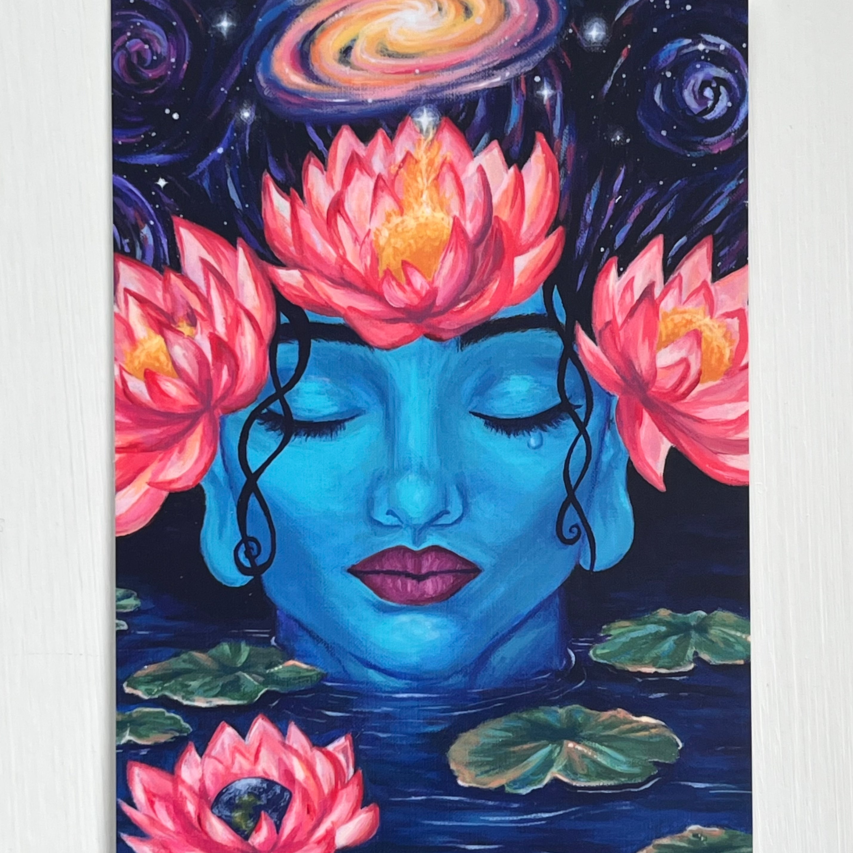 A postcard depicting artwork of the goddess Gaia, whose head is adorned with three large pink lotuses. She is surrounded by lilypads and a lotus that contains the planet Earth. Above her head, a galaxy swirls.