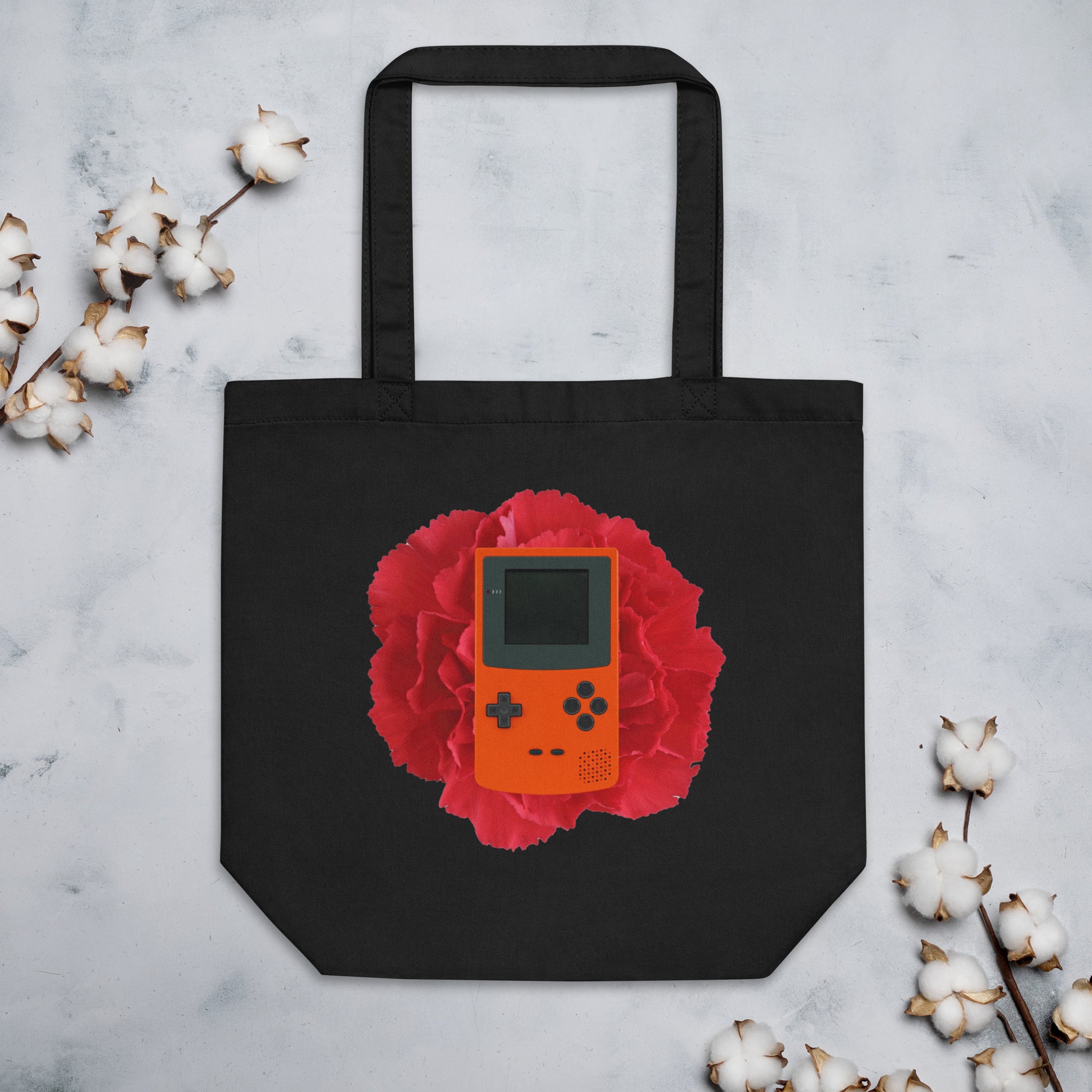 Black cotton tote bag featuring a design of an orange retro handheld game console (Gameboy) centered on a vibrant red flower. The tote bag is displayed against a light background with cotton stems around it.