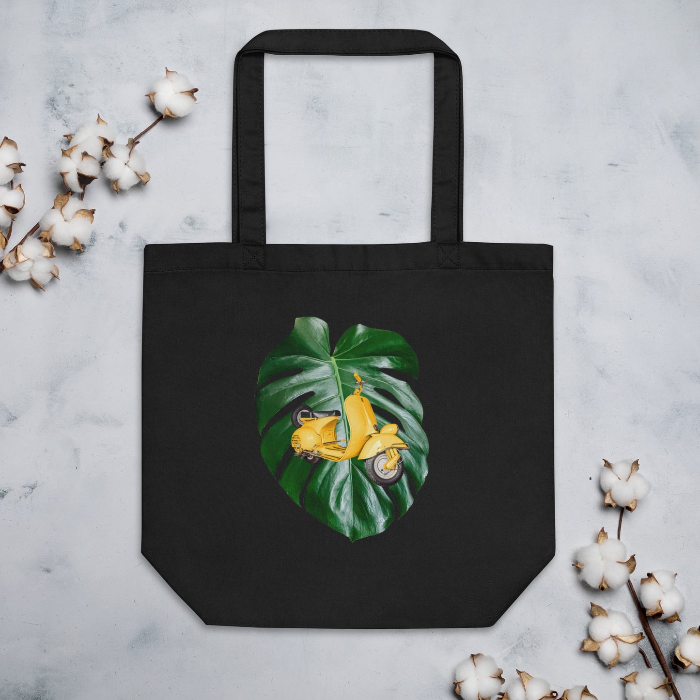 Black cotton tote bag featuring a design of a yellow scooter placed over a large green monstera leaf. The scooter is slightly angled to the right. The tote bag is displayed against a light background with cotton stems around it.