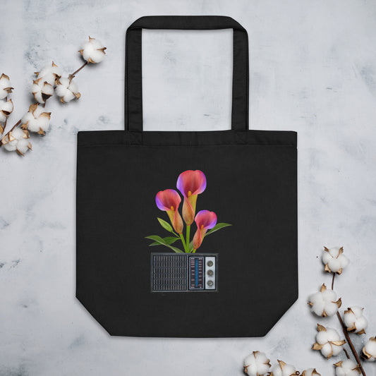 A black cotton tote bag featuring a graphic of pink and purple calla lilies growing out of a vintage radio. The bag is positioned on a marble surface, with sprigs of cotton flowers on both sides.