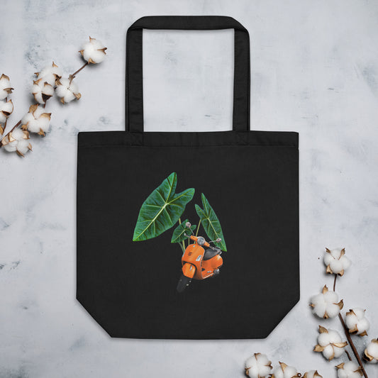 Black cotton tote bag featuring a design of an orange scooter overlaid on green tropical leaves (alocasias). The scooter is facing forward. The tote bag is displayed against a light background with cotton stems around it.