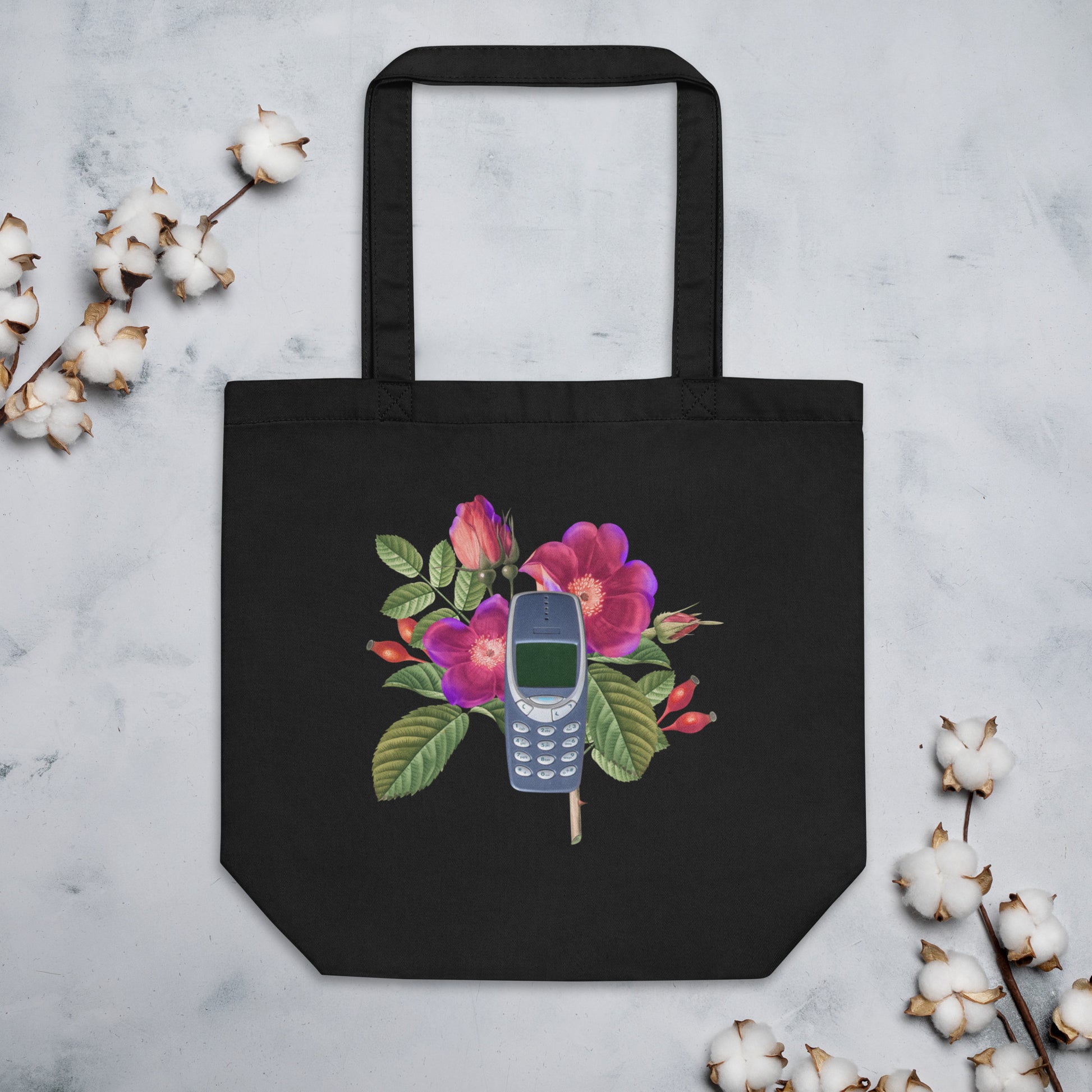 A black tote bag featuring a design of a vintage Nokia 3310 cellphone nestled among colorful flowers. The flowers include purple and pink blooms with green leaves.