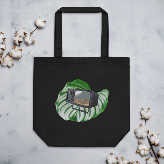 A black cotton tote bag featuring a graphic of a green monstera leaf with a vintage black television set placed in the center. The television screen displays a scenic desert landscape. The bag is positioned on a marble surface, with sprigs of cotton flowers on both sides.