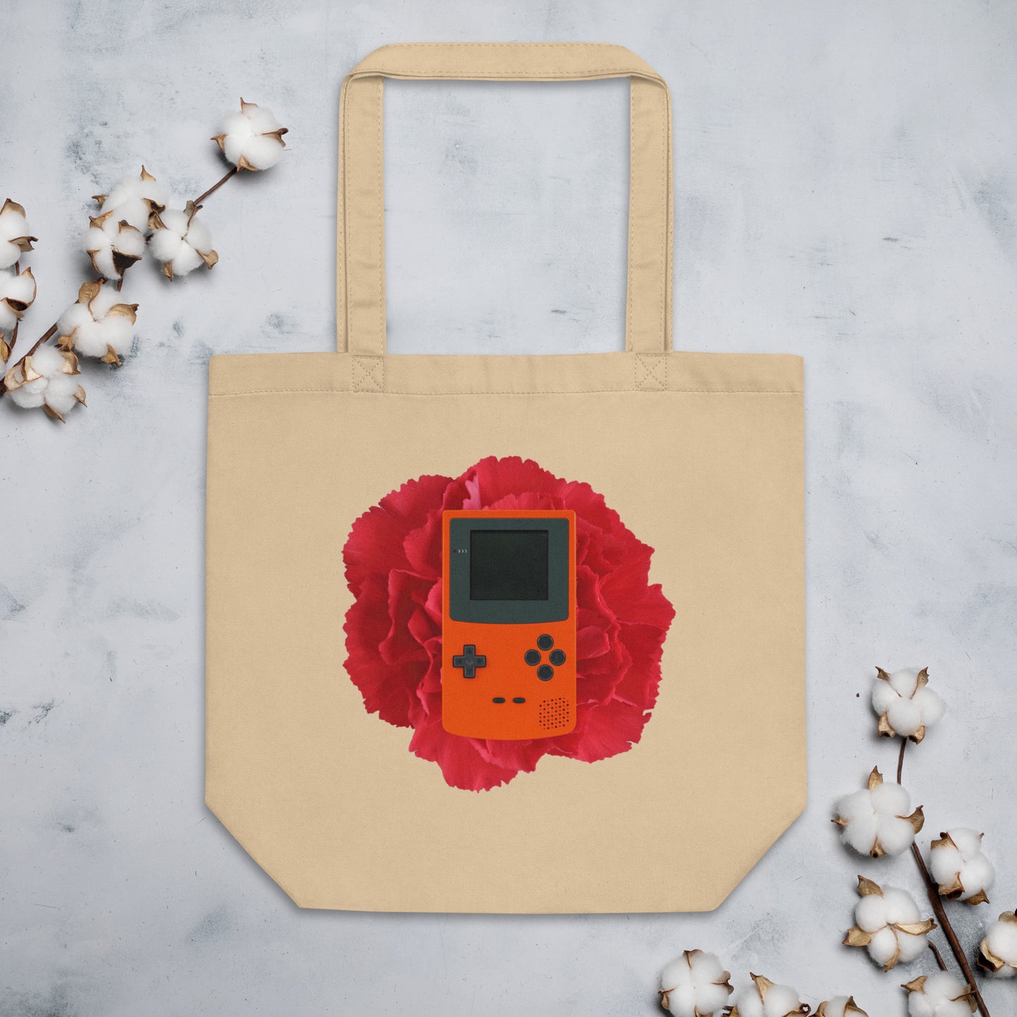 Beige cotton tote bag featuring a design of an orange retro handheld game console (Gameboy) centered on a vibrant red flower. The tote bag is displayed against a light background with cotton stems around it.