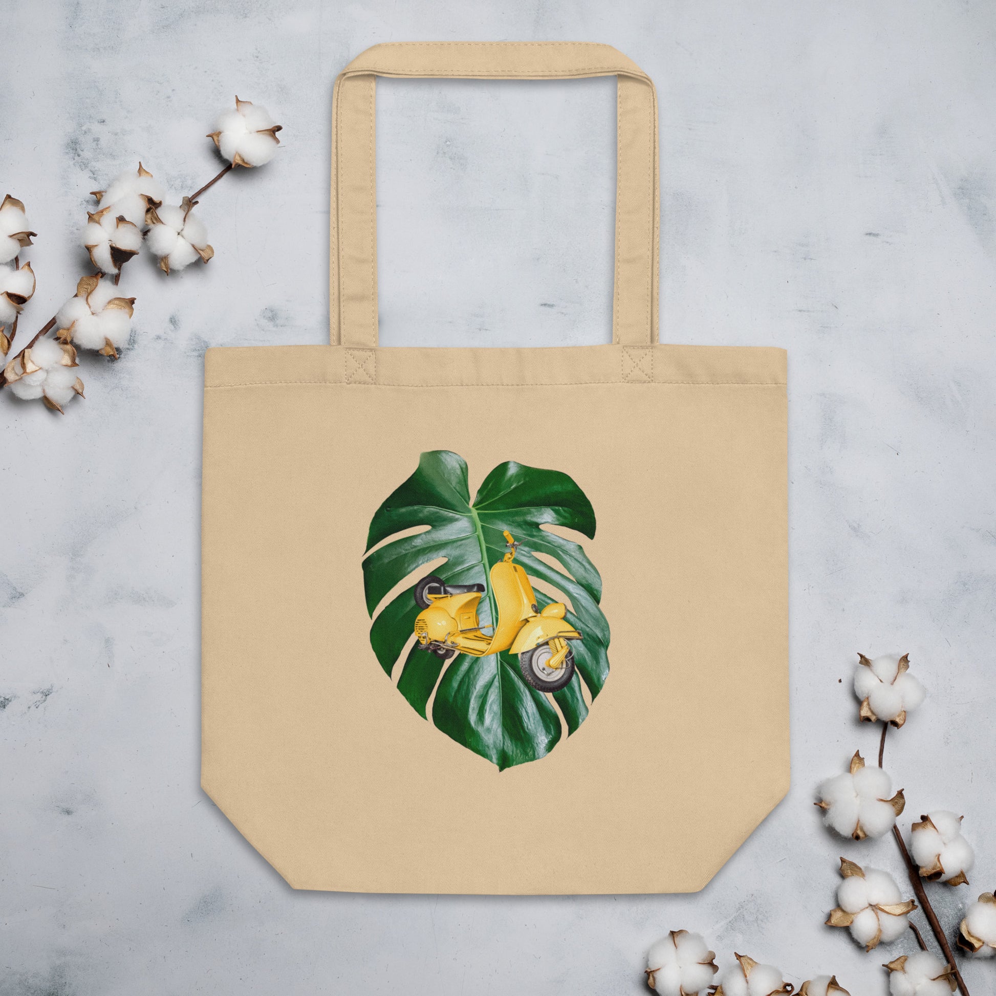 Beige cotton tote bag featuring a design of a yellow scooter placed over a large green monstera leaf. The scooter is slightly angled to the right. The tote bag is displayed against a light background with cotton stems around it.