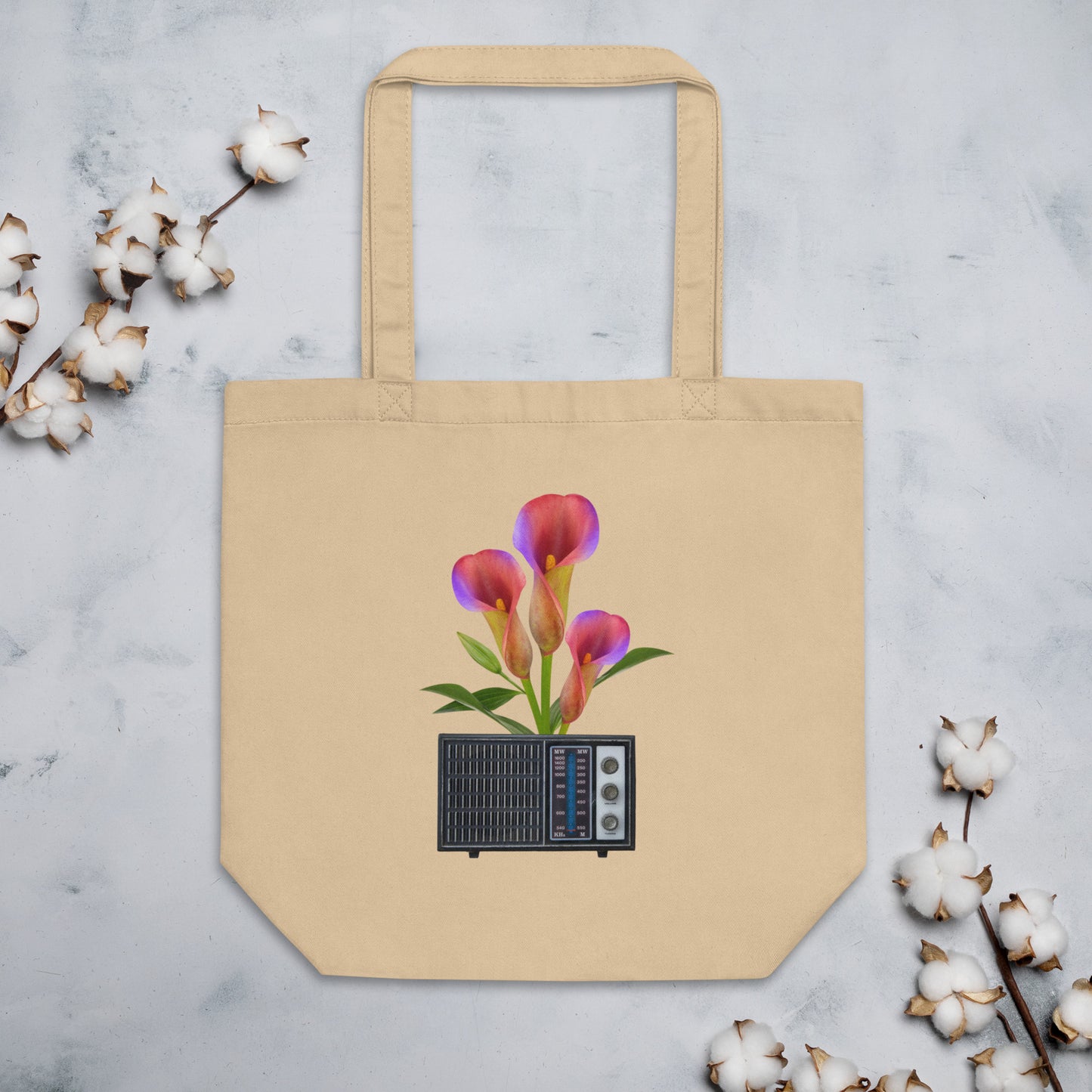 A beige cotton tote bag featuring a graphic of pink and purple calla lilies growing out of a vintage radio. The bag is positioned on a marble surface, with sprigs of cotton flowers on both sides.