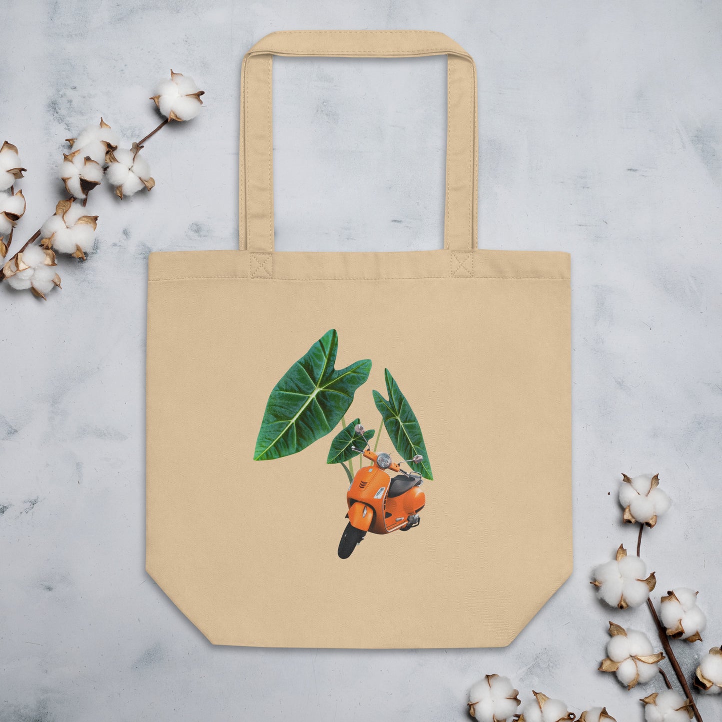 Beige cotton tote bag featuring a design of an orange scooter overlaid on green tropical leaves (alocasias). The scooter is facing forward. The tote bag is displayed against a light background with cotton stems around it.