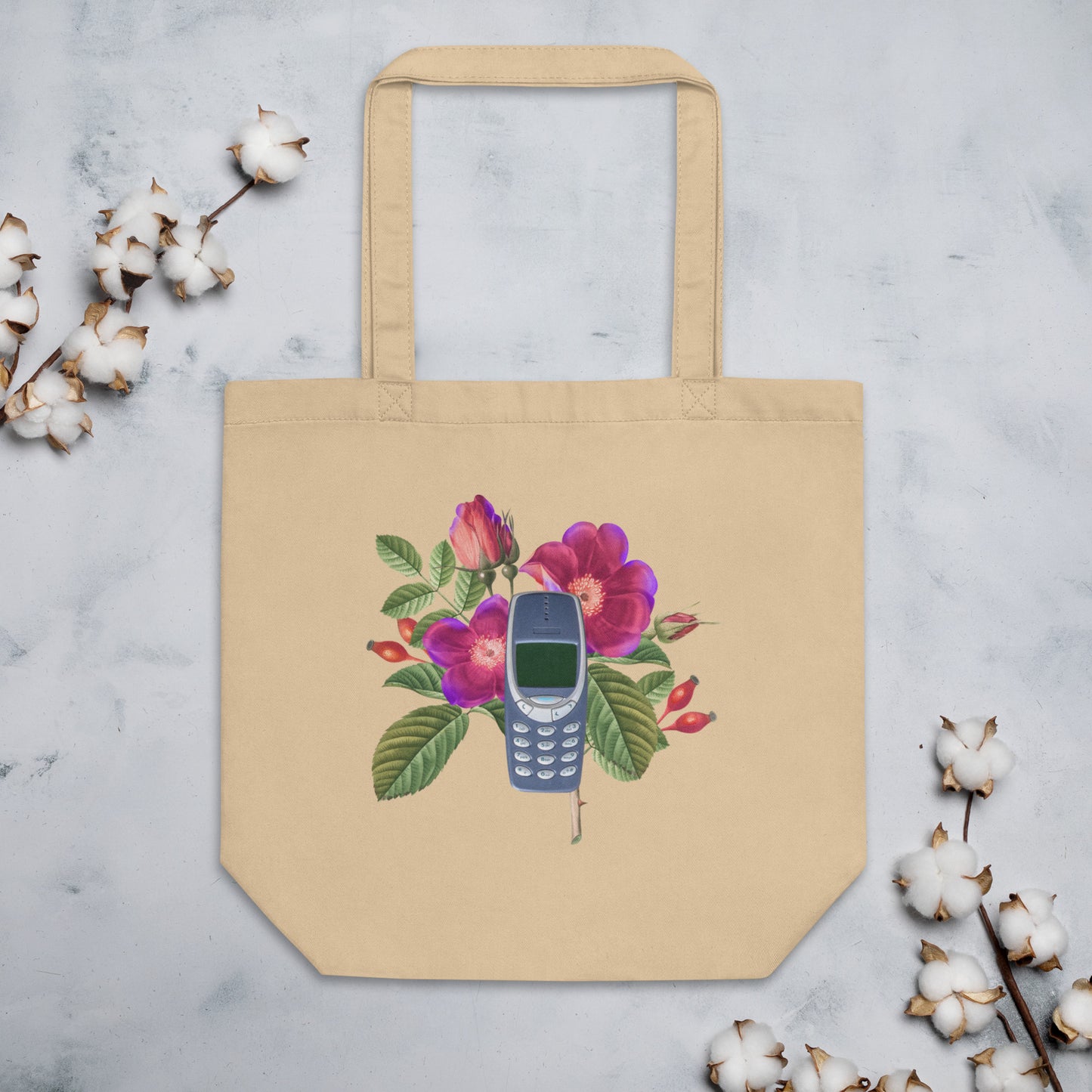 A tan tote bag featuring a design of a vintage Nokia 3310 cellphone nestled among colorful flowers. The flowers include purple and pink blooms with green leaves.