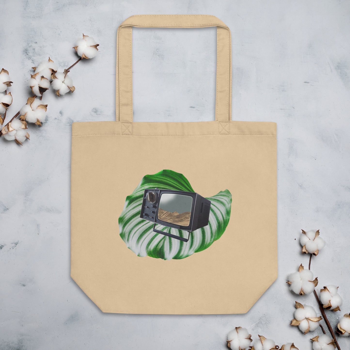 A beige cotton tote bag featuring a graphic of a green monstera leaf with a vintage black television set placed in the center. The television screen displays a scenic desert landscape. The bag is positioned on a marble surface, with sprigs of cotton flowers on both sides.