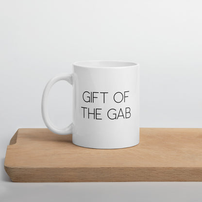 A glossy white ceramic mug with thin, all-caps black sans serif font that reads 'GIFT OF THE GAB', positioned on a wooden cutting board against a neutral background.