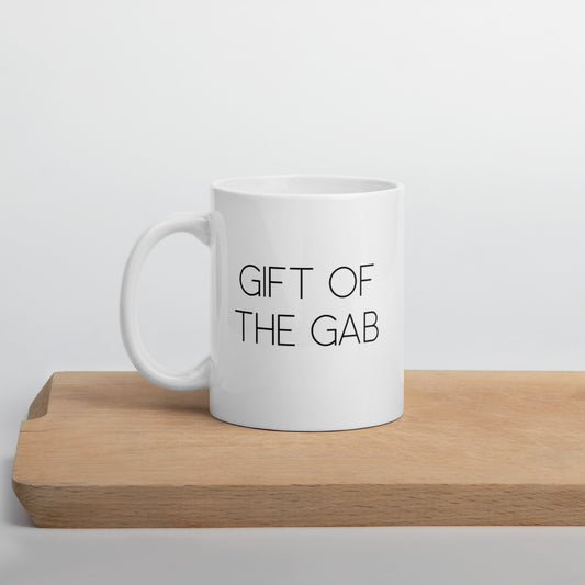 A glossy white ceramic mug with thin, all-caps black sans serif font that reads 'GIFT OF THE GAB', positioned on a wooden cutting board against a neutral background.