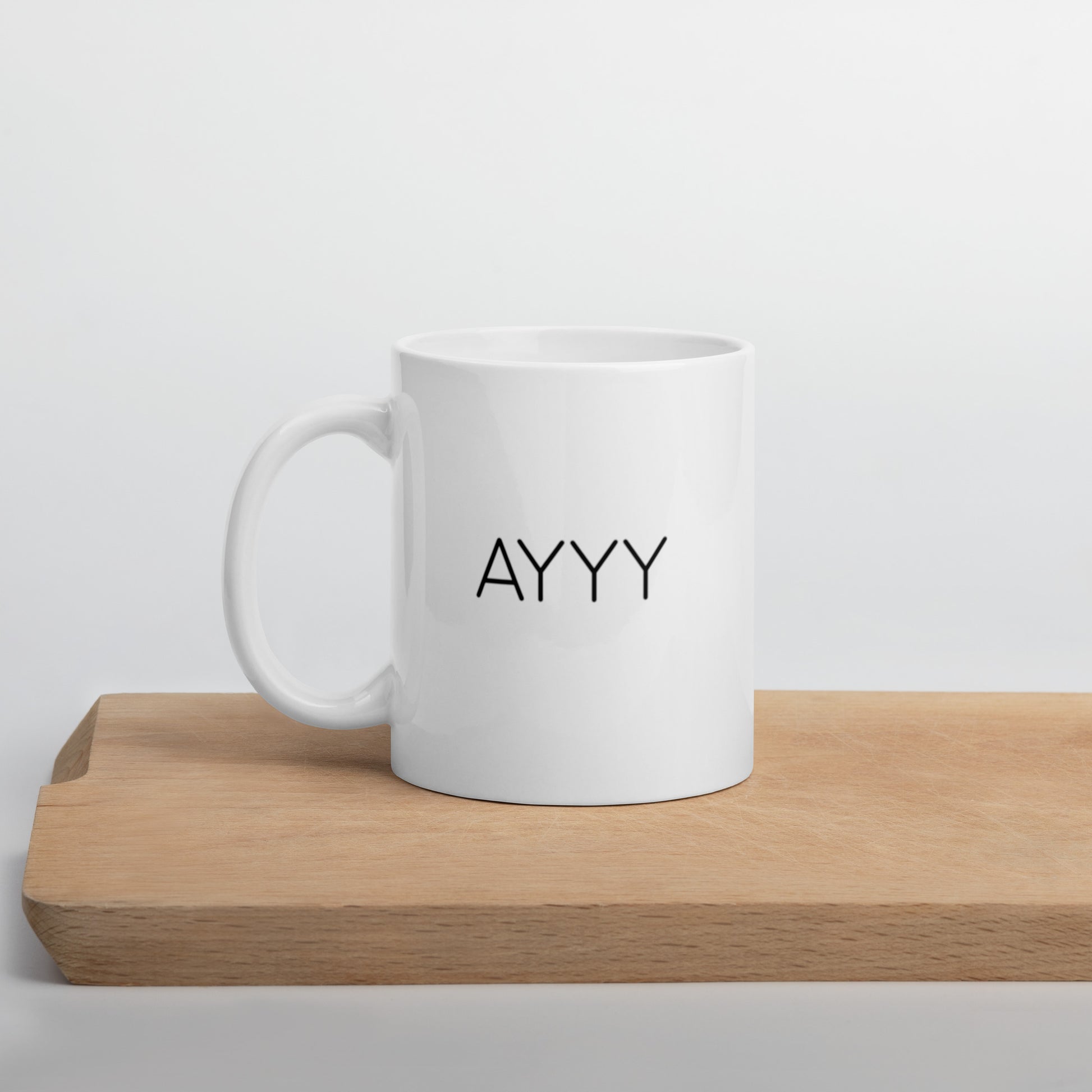 A glossy white ceramic mug with all-caps thin black text that reads 'AYYY', positioned on a wooden cutting board against a neutral background.