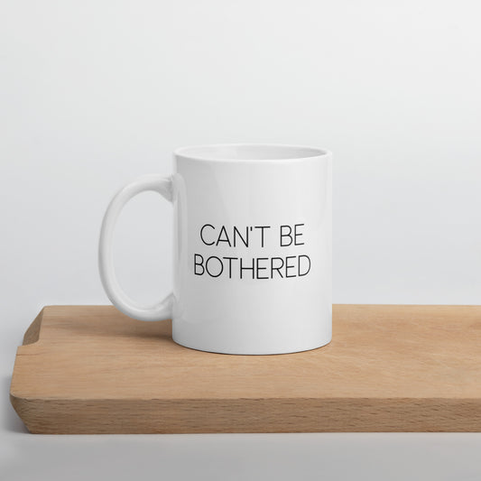 A glossy white ceramic mug with thin uppercase black text that reads 'CAN'T BE BOTHERED', positioned on a wooden cutting board against a neutral background.