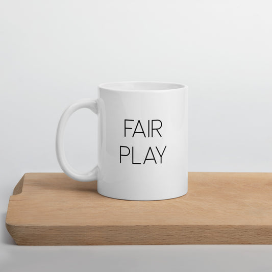 A glossy white ceramic mug with thin, all-caps black sans serif font that reads 'FAIR PLAY', positioned on a wooden cutting board against a neutral background.