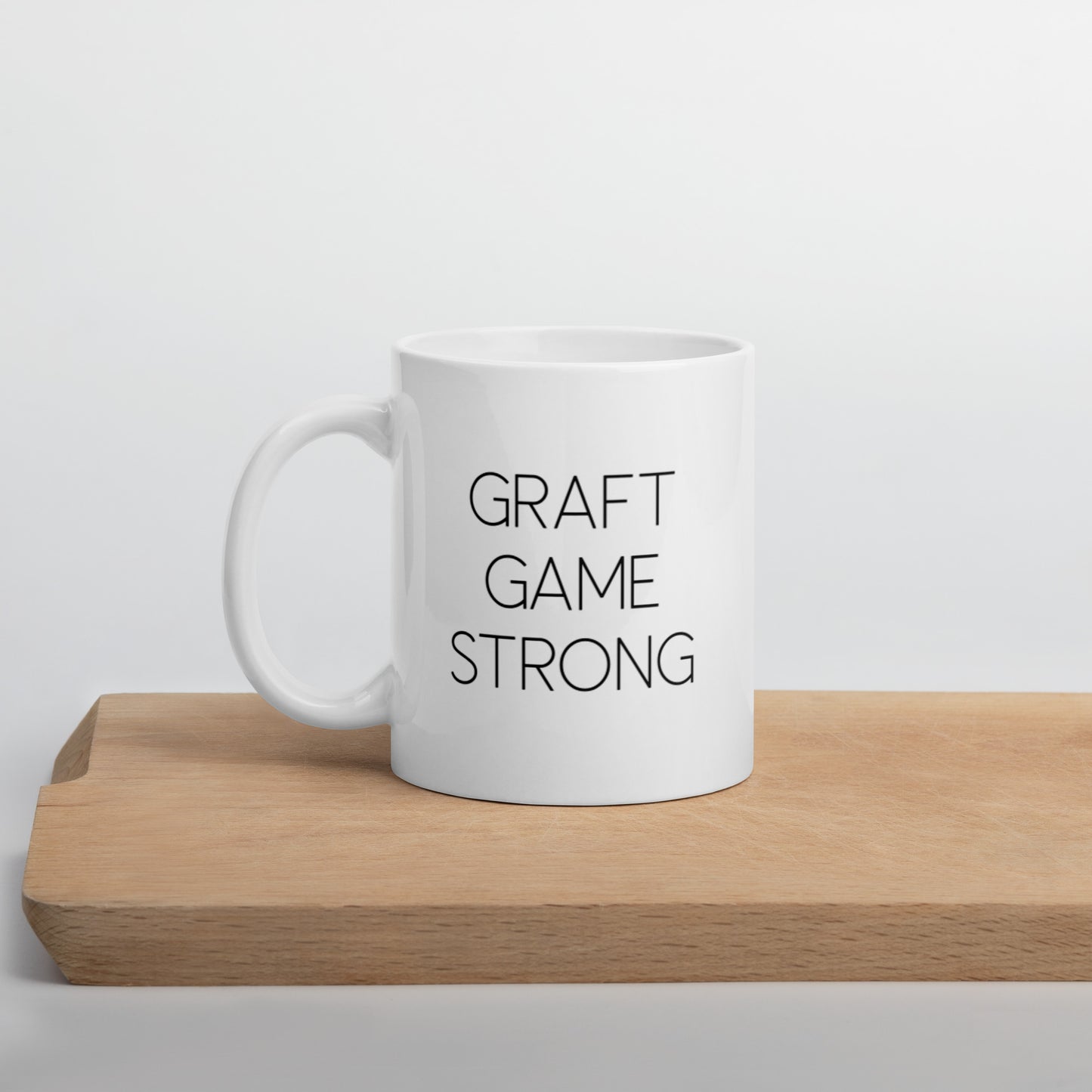 A glossy white ceramic mug with thin, all-caps black sans serif font that reads 'GRAFT GAME STRONG', positioned on a wooden cutting board against a neutral background.