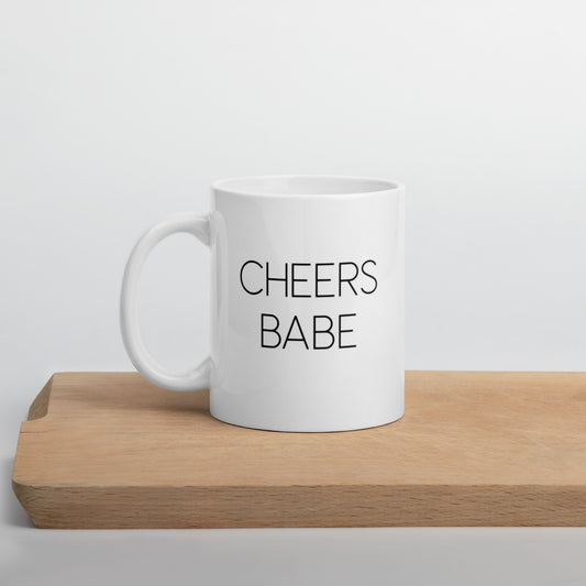 A glossy white ceramic mug with thin black all-caps text that reads 'CHEERS BABE', positioned on a wooden cutting board against a neutral background.