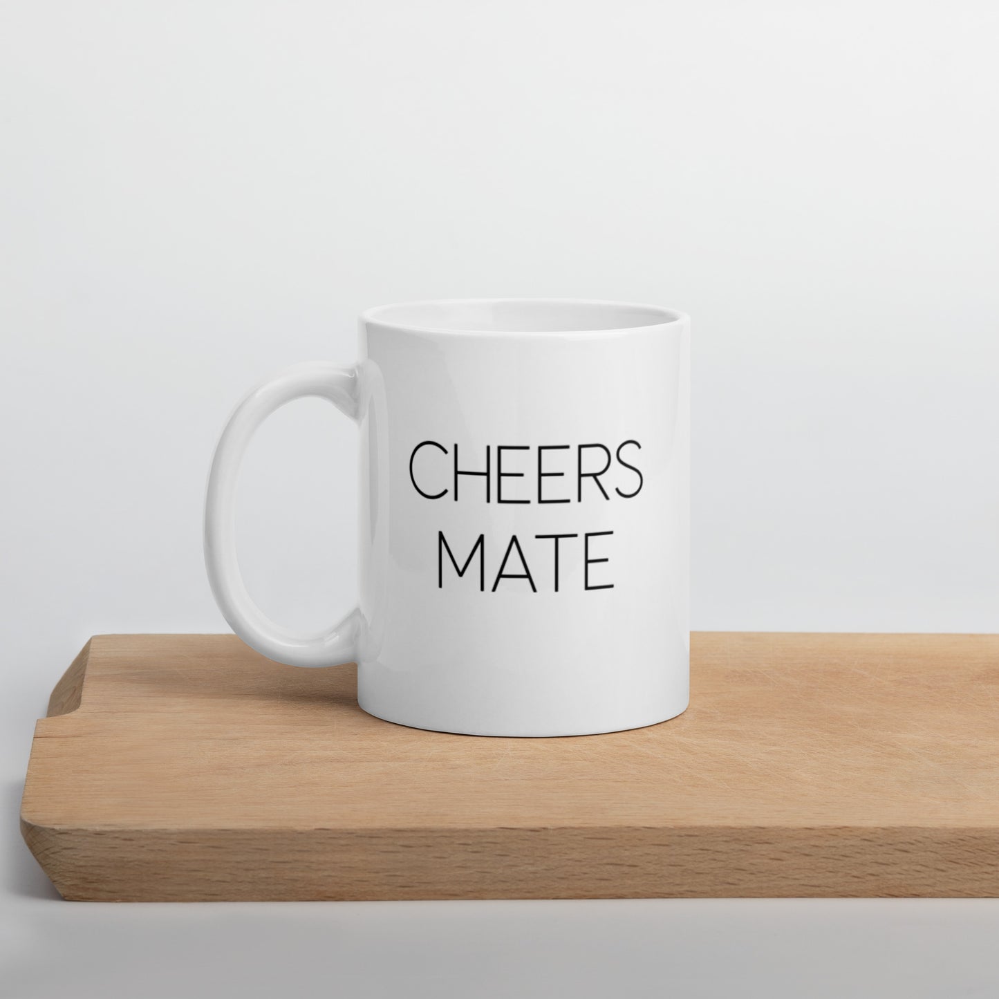 A glossy white ceramic mug with thin black all-caps text that reads 'CHEERS MATE', positioned on a wooden cutting board against a neutral background.