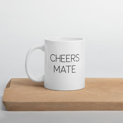 A glossy white ceramic mug with thin black all-caps text that reads 'CHEERS MATE', positioned on a wooden cutting board against a neutral background.