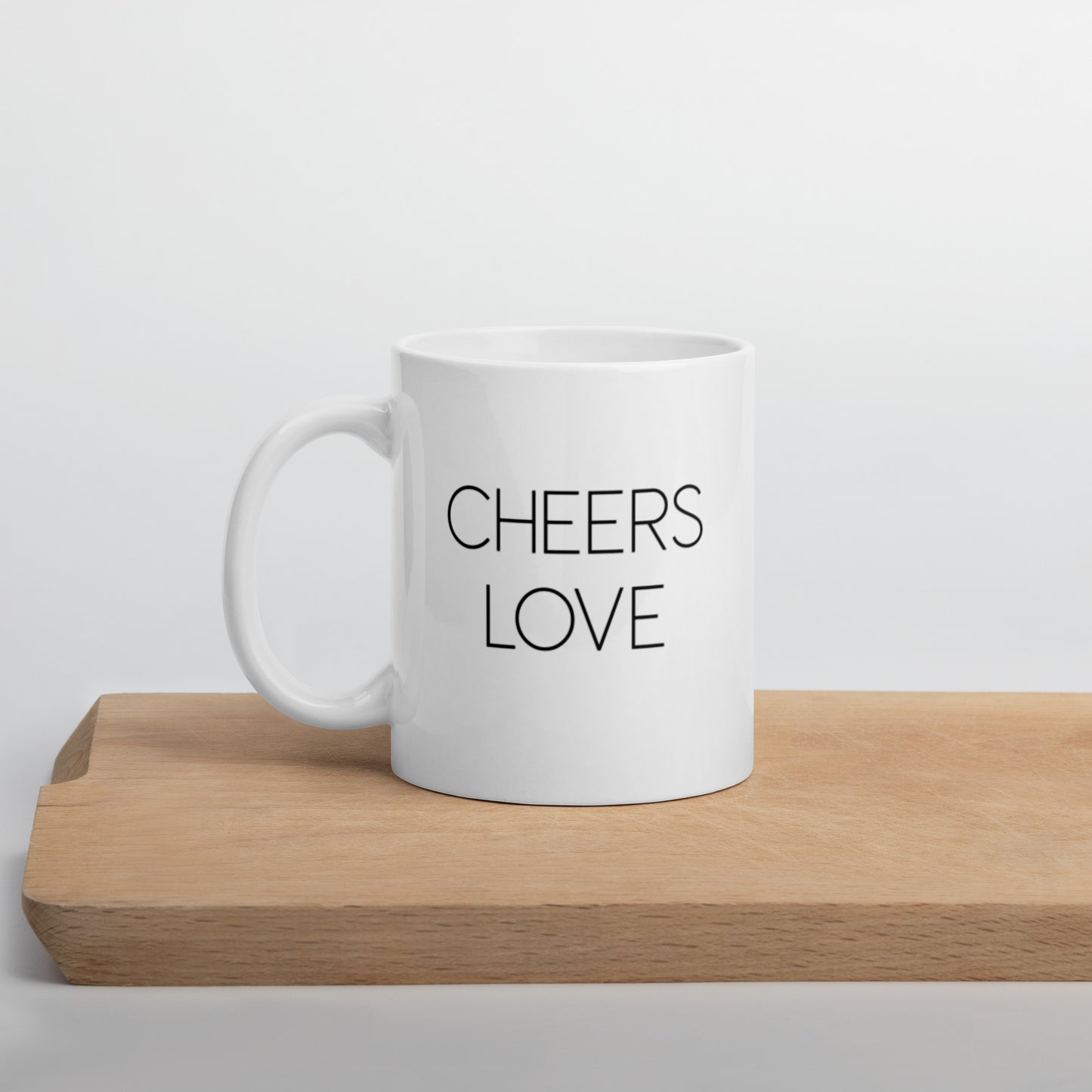 A glossy white ceramic mug with thin black all-caps text that reads 'CHEERS LOVE', positioned on a wooden cutting board against a neutral background.