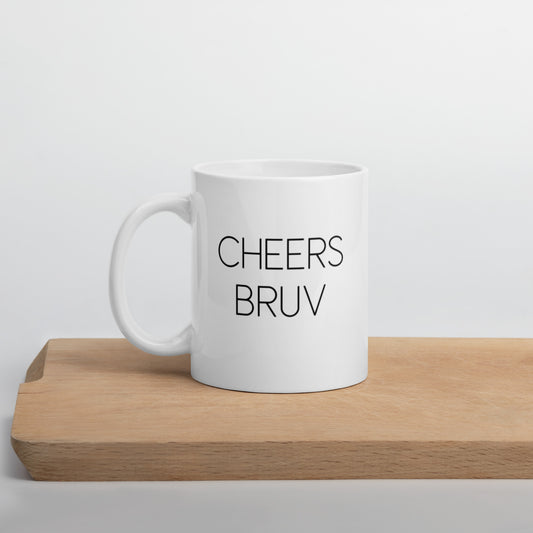 A glossy white ceramic mug with thin black all-caps text that reads 'CHEERS BRUV', positioned on a wooden cutting board against a neutral background.