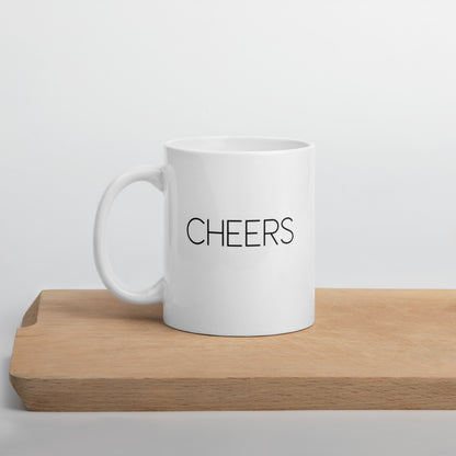 A glossy white ceramic mug with thin black all-caps text that reads 'CHEERS', positioned on a wooden cutting board against a neutral background.