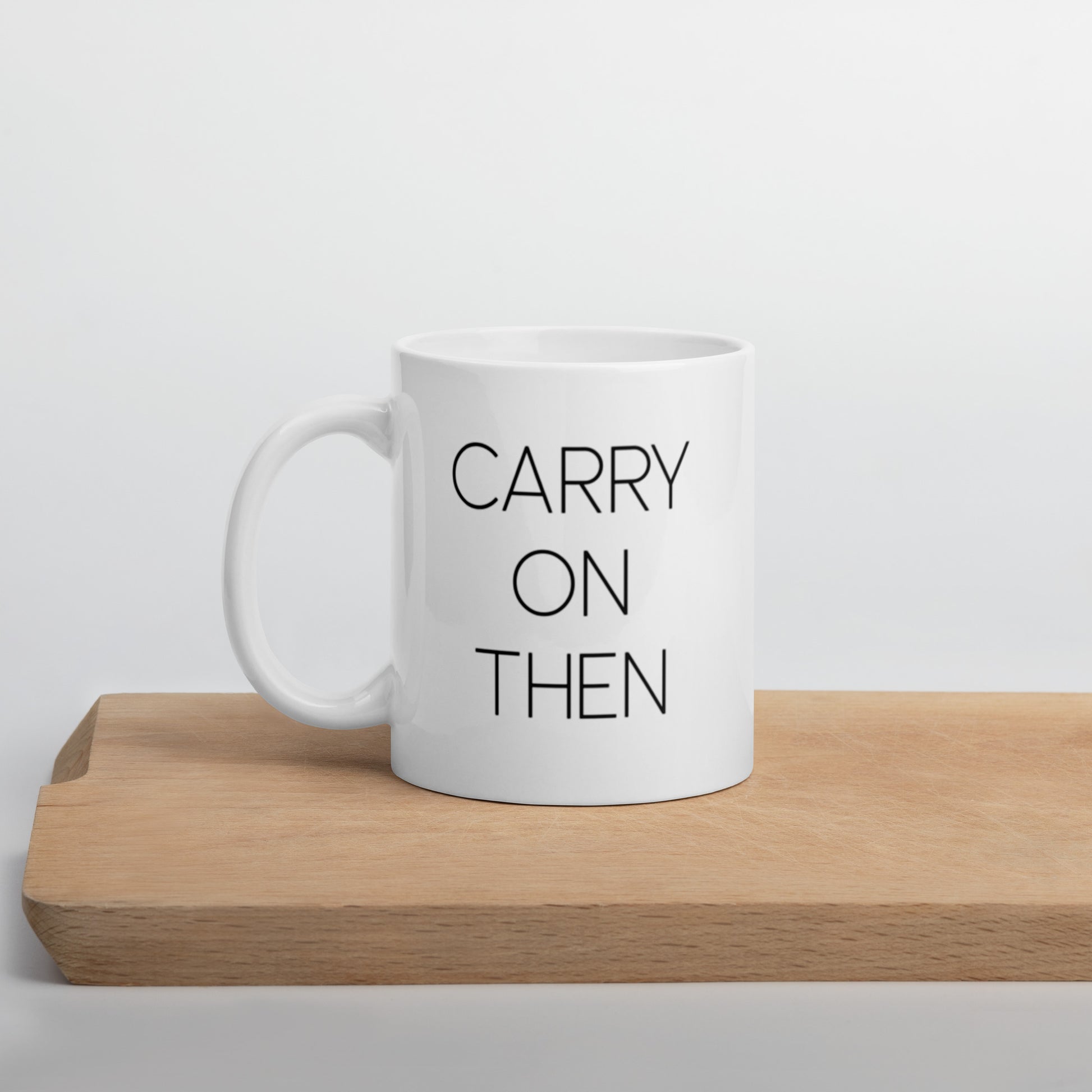 A glossy white ceramic mug with thin black all-caps text that reads 'CARRY ON THEN', positioned on a wooden cutting board against a neutral background.