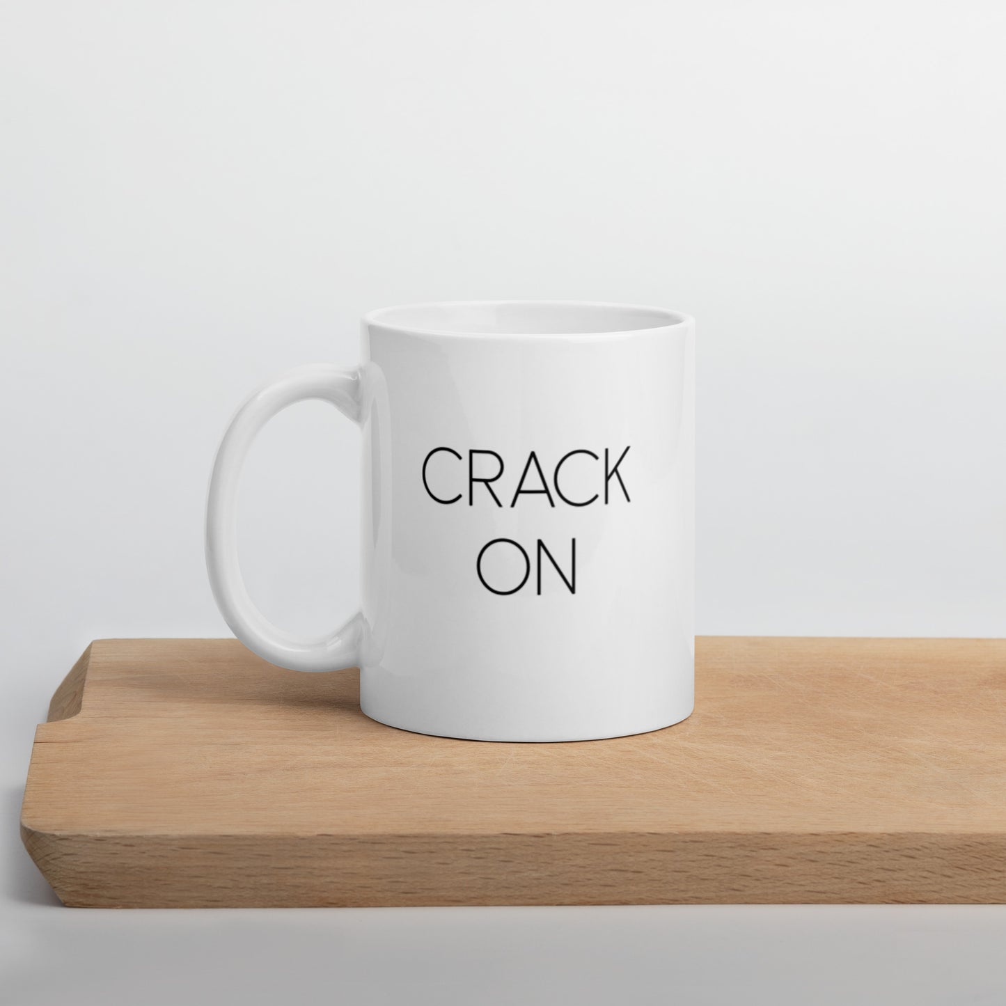 A glossy white ceramic mug with thin black all-caps text that reads 'CRACK ON', positioned on a wooden cutting board against a neutral background.