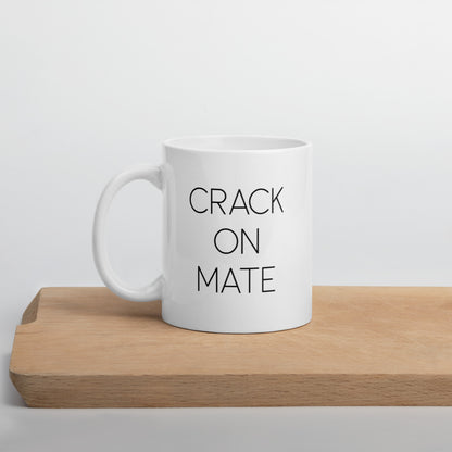 A glossy white ceramic mug with thin black all-caps text that reads 'CRACK ON MATE', positioned on a wooden cutting board against a neutral background.