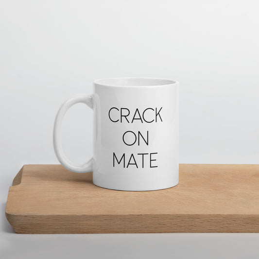 A glossy white ceramic mug with thin black all-caps text that reads 'CRACK ON MATE', positioned on a wooden cutting board against a neutral background.