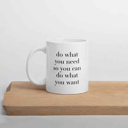 A glossy white ceramic mug with lowercase black serif font that reads 'do what you need so you can do what you want', positioned on a wooden cutting board against a neutral background.