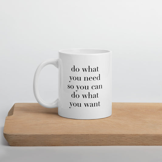 A glossy white ceramic mug with lowercase black serif font that reads 'do what you need so you can do what you want', positioned on a wooden cutting board against a neutral background.