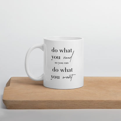 A glossy white ceramic mug with lowercase black serif font that reads 'do what you need so you can do what you want' ('so you can' is in smaller font, and 'need' and 'want' are in script to provide contrast with the serif font), positioned on a wooden cutting board against a neutral background.