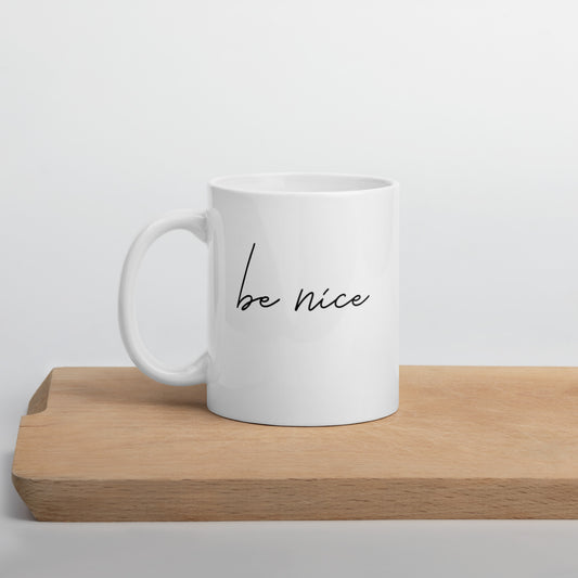 A glossy white ceramic mug with black script text that reads 'be nice', positioned on a wooden cutting board against a neutral background.