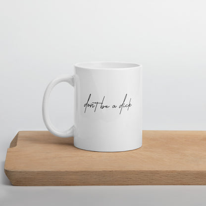 A glossy white ceramic mug with lowercase script font that reads 'don't be a dick', positioned on a wooden cutting board against a neutral background.