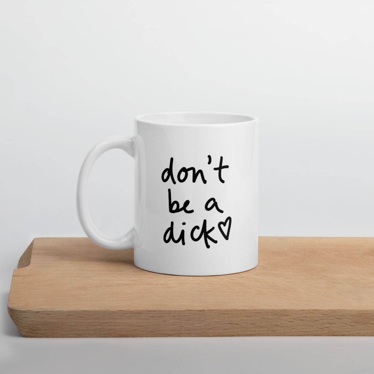 A glossy white ceramic mug with bold handwritten-style font that reads 'don't be a dick [ hand-drawn heart ]', positioned on a wooden cutting board against a neutral background.