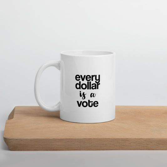 A glossy white ceramic mug with bold, lowercase black sans serif font that reads 'every dollar is a vote', with 'is a' in black handwritten script font, positioned on a wooden cutting board against a neutral background.