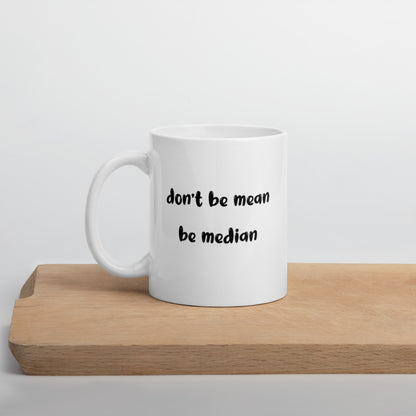 A glossy white ceramic mug with bold, lowercase black bubble font that reads 'don't be mean be median', positioned on a wooden cutting board against a neutral background.