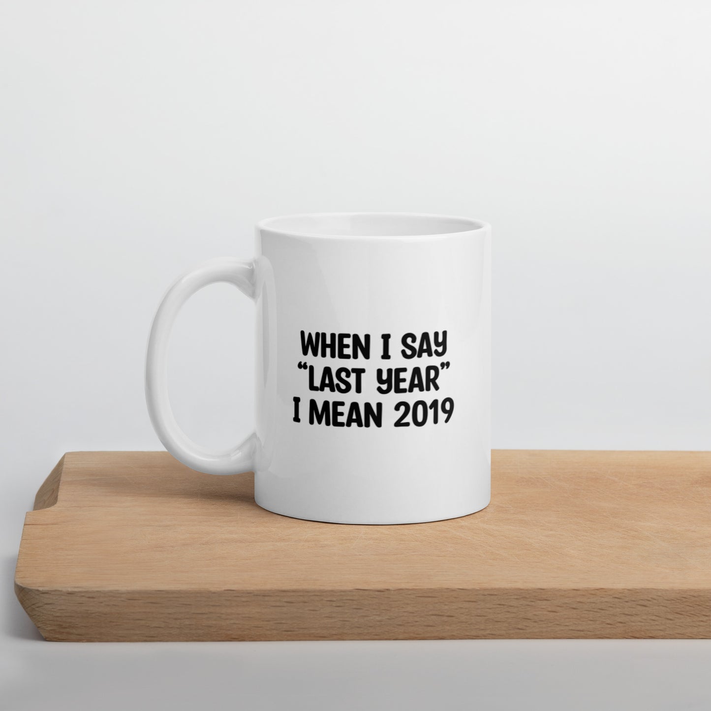 A glossy white ceramic mug with the text 'WHEN I SAY 'LAST YEAR' I MEAN 2019' in black bold uppercase font, positioned on a wooden cutting board against a neutral background.