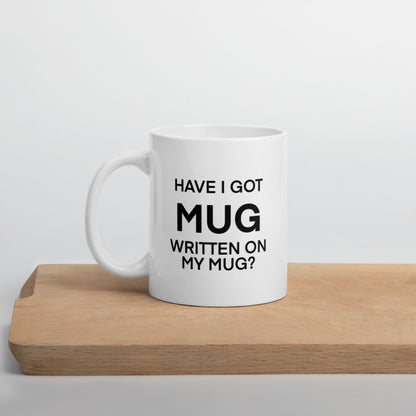 A glossy white ceramic mug with black bolded sans serif text that reads 'HAVE I GOT MUG WRITTEN ON MY MUG?', where the first 'MUG' is in bigger font size, positioned on a wooden cutting board against a neutral background.