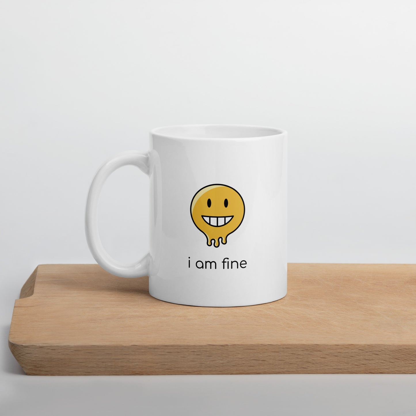 A glossy white ceramic mug with a graphic of a yellow smiley face (grinning) and melting at the bottom, accompanied by the caption 'i am fine' in small black sans serif text, positioned on a wooden cutting board against a neutral background.