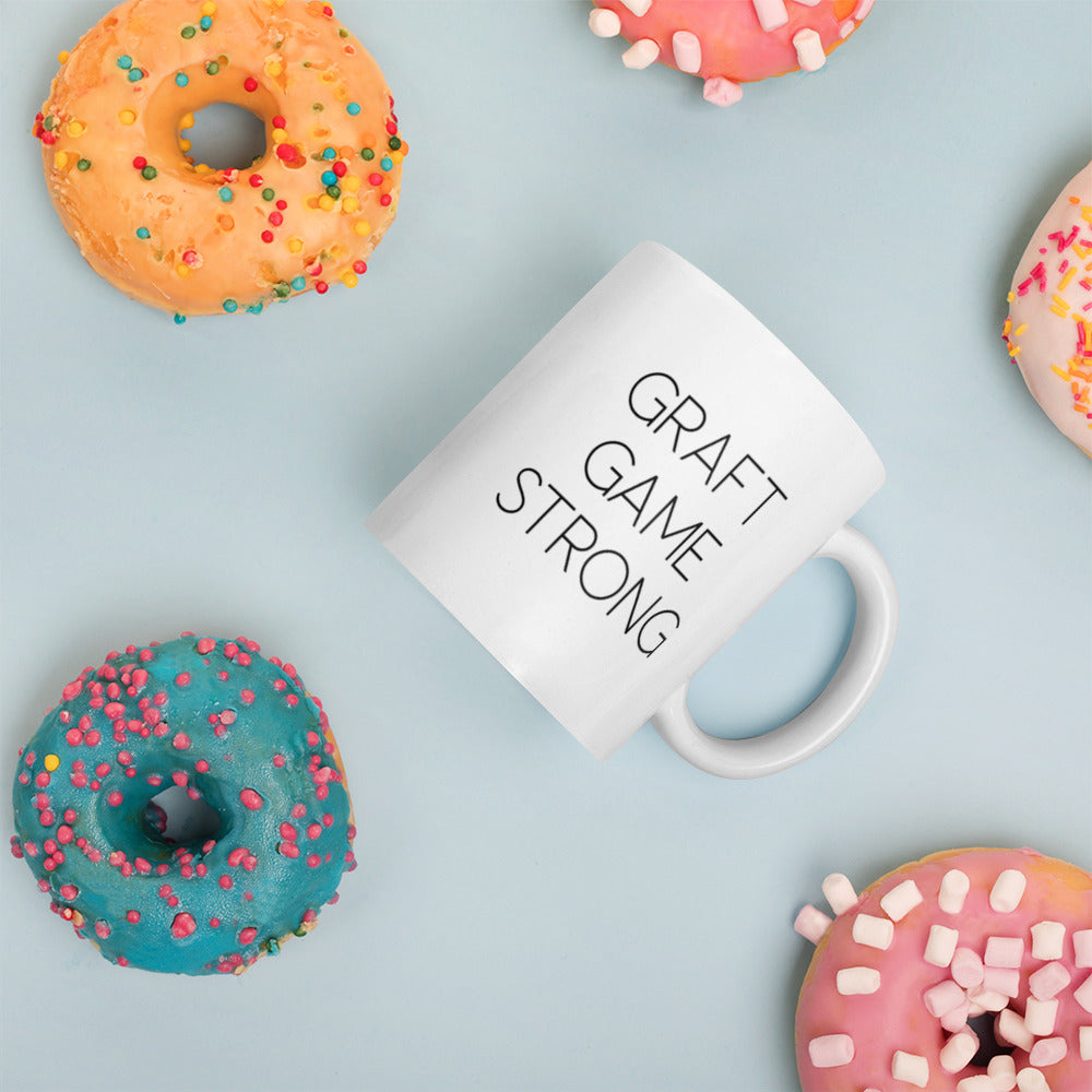 A glossy white ceramic mug with thin, all-caps black sans serif font that reads 'GRAFT GAME STRONG', placed on its side against a pastel light blue background. Colorful doughnuts are scattered around the mug, adding a playful touch to the scene.