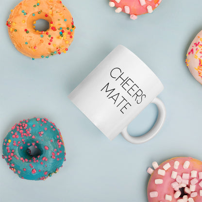 A glossy white ceramic mug with thin black all-caps text that reads 'CHEERS MATE', placed on its side against a pastel light blue background. Colorful doughnuts are scattered around the mug, adding a playful touch to the scene.