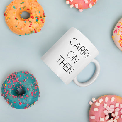 A glossy white ceramic mug with thin black all-caps text that reads 'CARRY ON THEN', placed on its side against a pastel light blue background. Colorful doughnuts are scattered around the mug, adding a playful touch to the scene.