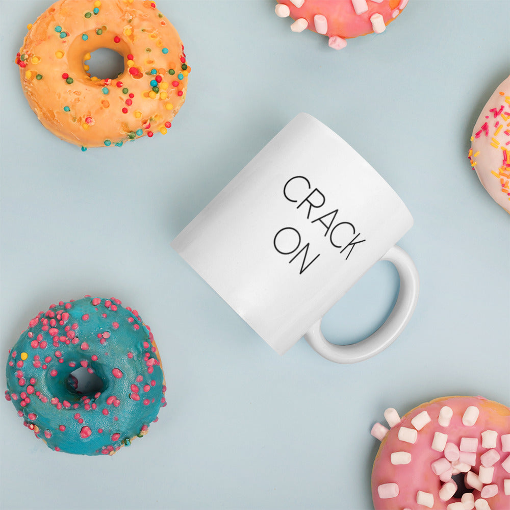 A glossy white ceramic mug with thin black all-caps text that reads 'CRACK ON', placed on its side against a pastel light blue background. Colorful doughnuts are scattered around the mug, adding a playful touch to the scene.