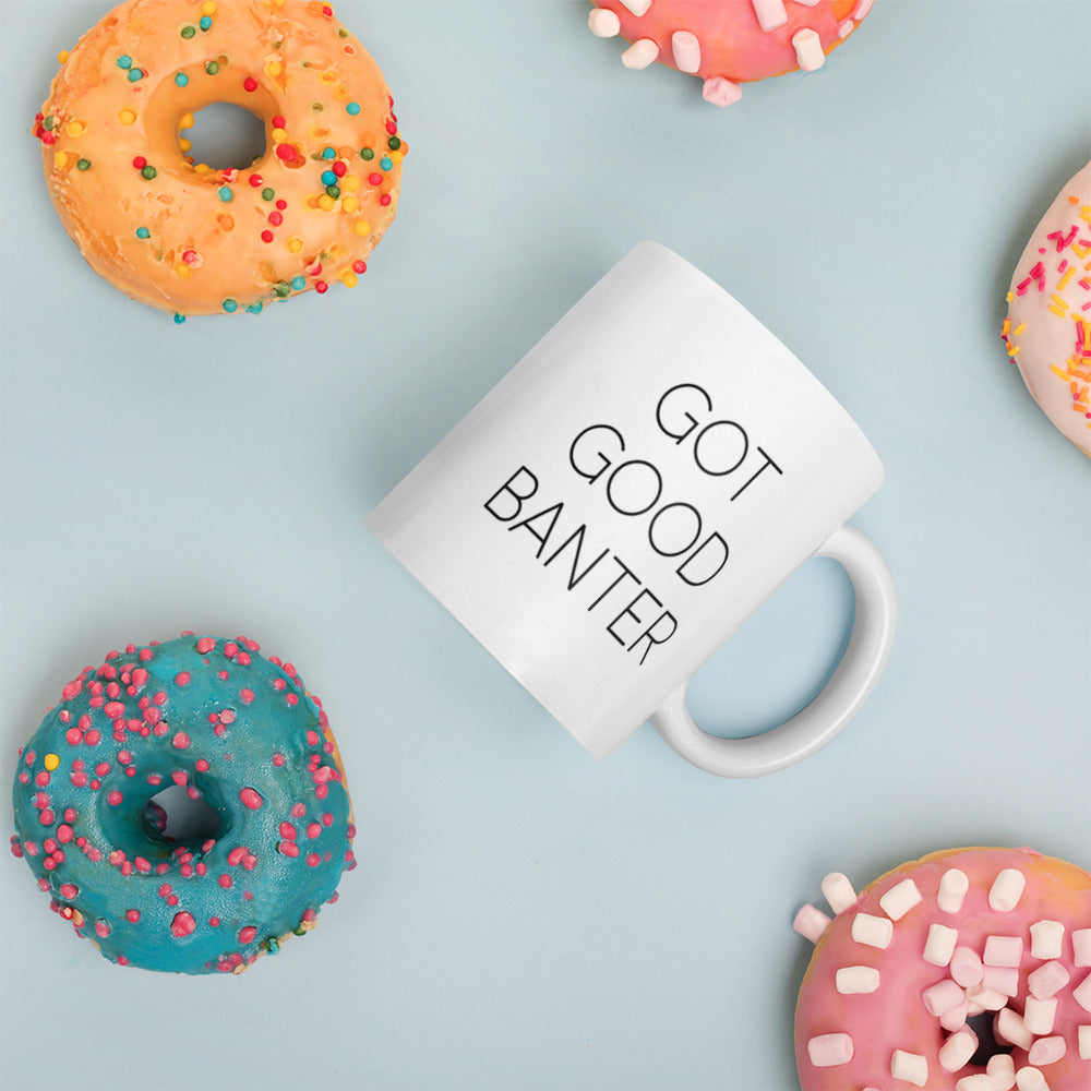 A glossy white ceramic mug with thin, all-caps black sans serif font that reads 'GOT GOOD BANTER', placed on its side against a pastel light blue background. Colorful doughnuts are scattered around the mug, adding a playful touch to the scene.