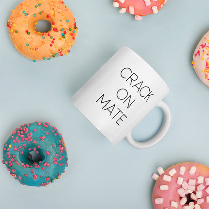 A glossy white ceramic mug with thin black all-caps text that reads 'CRACK ON MATE', placed on its side against a pastel light blue background. Colorful doughnuts are scattered around the mug, adding a playful touch to the scene.