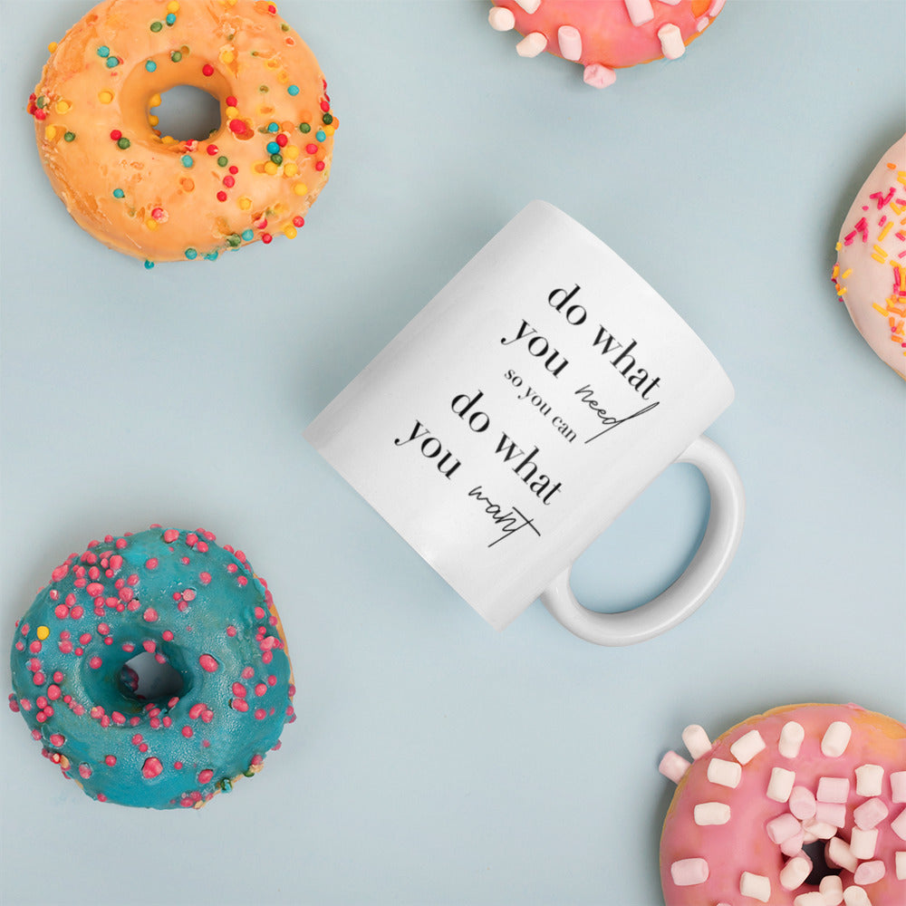 A glossy white ceramic mug with lowercase black serif font that reads 'do what you need so you can do what you want' ('so you can' is in smaller font, and 'need' and 'want' are in script to provide contrast with the serif font), placed on its side against a pastel light blue background. Colorful doughnuts are scattered around the mug, adding a playful touch to the scene.