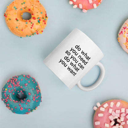 A glossy white ceramic mug with lowercase bolded black sans serif font that reads 'do what you need so you can do what you want', placed on its side against a pastel light blue background. Colorful doughnuts are scattered around the mug, adding a playful touch to the scene.