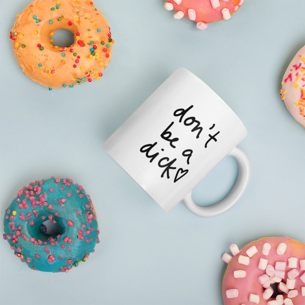 A glossy white ceramic mug with bold handwritten-style font that reads 'don't be a dick [ hand-drawn heart ]', placed on its side against a pastel light blue background. Colorful doughnuts are scattered around the mug, adding a playful touch to the scene.