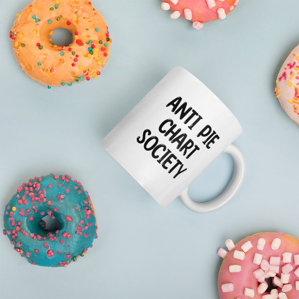 A glossy white ceramic mug with all-uppercase black bubble text that reads 'ANTI PIE CHART SOCIETY', placed on its side against a pastel light blue background. Colorful doughnuts are scattered around the mug, adding a playful touch to the scene.